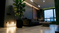 Modern luxury living room with neon hello sign, dark interior design, large plant, floor-to-ceiling window, and city view at dusk.