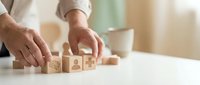 Businessman hands arranging wooden blocks with business icons on white desk in modern office environment for strategic planning and goal setting.