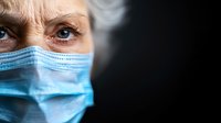 Senior Caucasian woman wearing protective blue surgical face mask with focused gaze and wrinkled eyes against dark background, healthcare concept.