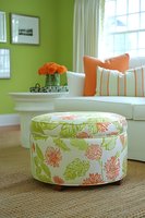 Modern living room interior featuring floral ottoman in coral and green pattern, white sofa with orange accent pillows against lime green walls and natural light.