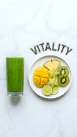 Green smoothie in a tall glass beside a plate of tropical fruits with vitality text made of chia seeds on marble background.