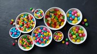 Colorful assorted candies and sweets arranged in white bowls on dark slate background. Variety of gummy bears, chocolate balls and rainbow treats for party.