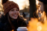 Young Asian woman in winter attire smiling while holding coffee cup outdoors during golden hour sunset, wearing knit beanie and fur-trimmed coat.
