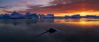 Icebergs floating in arctic waters during golden hour sunset with dramatic sky and ice formations reflecting in calm ocean surface.