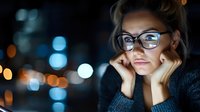 Thoughtful young woman with glasses resting chin on hands against blurred city lights at night, conveying contemplation and solitude.