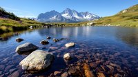 Tranquil alpine lake with scattered boulders reflecting snow-capped mountain peaks, clear water reveals rocky bottom amid green meadows and pristine wilderness landscape.