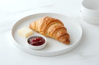 Fresh baked croissant served on white ceramic plate with butter and raspberry jam, minimalist breakfast composition on light background.