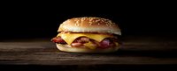 Bacon cheeseburger with melted cheese and crispy strips on sesame bun against dark background on rustic wooden table, dramatic lighting creates appetizing mood.
