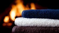 Stack of folded soft cotton towels in navy blue, white and brown colors against blurred fireplace background creating cozy spa atmosphere at home.