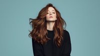 Young woman with flowing red hair wearing black sweater against turquoise background, eyes closed in serene expression. Professional beauty and fashion portrait.