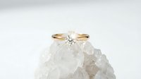 Elegant gold solitaire diamond engagement ring resting on a raw crystal rock, sparkling gemstone catching light on a soft white background.