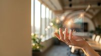 Woman's hand holding transparent glass panel with glowing light bulb icon against bright window and modern office interior background for innovation and technology concepts.