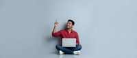 Man in red shirt sitting cross-legged with laptop, pointing upward against gray background. Suitable for business, technology, education, and online learning concepts.