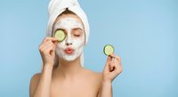 Young woman with white towel on head applying cucumber face mask for skincare routine and spa treatment at home on blue background.