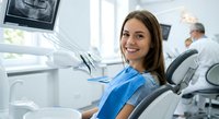 Young Caucasian woman patient smiling during dental examination in modern clinic with dentist working in background and x-ray monitor visible.