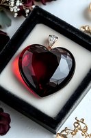 Deep red crystal heart pendant with silver bail in black velvet jewelry box, surrounded by romantic decor elements and rose petals on white surface.