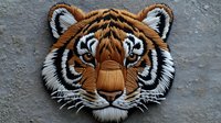 Detailed tiger head embroidery patch with orange, white and black threads on gray fabric background shows intricate needlework and textural elements.