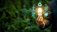 Businessman holding glowing lightbulb with green energy icons including recycling, wind turbine, solar panel and leaf symbols representing sustainable innovation.