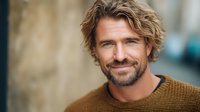 Caucasian male with shoulder-length blonde hair and facial hair wearing mustard knit sweater, smiling at camera outdoors for lifestyle and portrait use.
