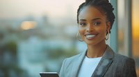 Young African American businesswoman with braided hair and bright smile wearing grey blazer and white top against blurred office background in warm evening light.