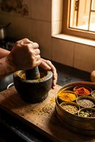 Elderly hands grinding spices in traditional mortar and pestle with colorful spice collection in rustic kitchen setting for authentic cooking preparation.