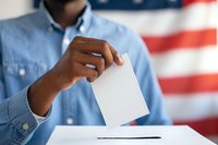 African American voter casting ballot at polling station with USA flag background, perfect for election coverage, democracy, and civic engagement themes.