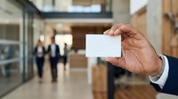 Mature male professional in business suit holding blank white business card in modern office lobby with colleagues in background, corporate networking and branding concept.