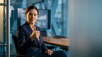 Mature woman in dark blazer giving thumbs up gesture in modern office environment with glass partitions and wooden desk.