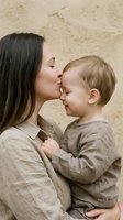 Young mother kissing toddler son forehead in tender moment. Caucasian woman with dark hair embracing blonde child in beige linen clothing against neutral background.