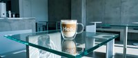 Glass cup of latte coffee with Hello text on foam, placed on a glass table in a modern minimalist interior with concrete walls.