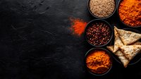 Vibrant Indian spices in black bowls with samosas on dark stone background, colorful cooking ingredients for authentic Asian cuisine preparation.