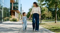 Young Caucasian mother and daughter walking hand in hand on sidewalk near modern home. Family bonding time outdoors in residential neighborhood setting.