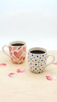 Two ceramic mugs filled with black coffee sit on a wooden surface with scattered flower petals, one featuring red hearts and the other with blue dots.