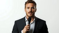 Confident male speaker in black suit and white shirt holding microphone against white background, professional business portrait for presentations and events.