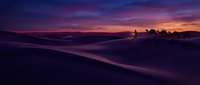 Silhouetted figures stand on a sandy beach at dusk, watching the dramatic purple and pink sunset reflect across calm ocean waters and distant coastline.