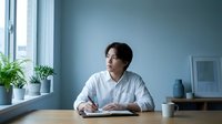 Young East Asian man in business casual attire sitting at desk with notebook, coffee cup, and potted plants by window in modern office environment for corporate, professional, and workplace concepts.