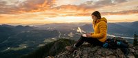 Woman in yellow jacket sitting on mountain rock with laptop, working remotely at sunset overlooking forested valley and distant peaks.