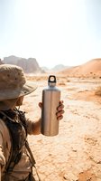 Traveler holding metal water bottle in arid desert landscape with mountains, wearing hat and backpack. Hydration and adventure concept for outdoor activities and travel campaigns.