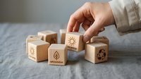 Hand placing wooden cube with sun icon among eco-friendly blocks featuring leaf and recycling symbols on gray surface for sustainable business concepts.