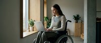 Woman with dark hair and light skin tone, appearing to be in her thirties, seated in a wheelchair while working on a laptop in a modern, minimalist office space with natural light and potted plants.