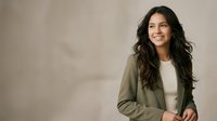 Young woman with long dark wavy hair and olive skin tone wearing casual olive green cardigan over white top, smiling confidently against neutral beige background. Professional portrait for business, lifestyle, and corporate use.