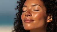 Young African American woman with curly hair enjoying sunlight with closed eyes and serene expression, glowing skin reflects golden hour lighting on beach background.
