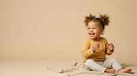 Cheerful toddler girl with curly brown hair wearing yellow shirt sitting on beige background, laughing and playing. For childcare, parenting, and family lifestyle content.