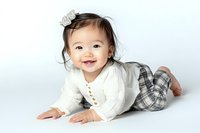 Cheerful Asian baby girl in white cardigan and plaid pants lying on stomach, smiling brightly with natural expression against light background.