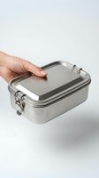 Hand holding a stainless steel lunch box with metal clasps on a light background, eco-friendly reusable food container.