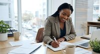 Professional African American businesswoman writing notes at modern office desk with laptop and coffee cup near window.