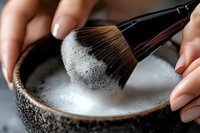 Makeup brush being cleaned in soapy water in ceramic bowl, close-up view showing foam and bristles. Professional beauty tool maintenance and hygiene routine.