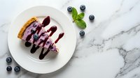 Blueberry cheesecake slice with berry sauce drizzle on white plate, fresh blueberries and mint garnish on marble surface for dessert menu photography.