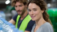 Young female warehouse worker smiling in distribution center with coworker in safety vest in background. Modern logistics facility environment shows professional workplace setting.