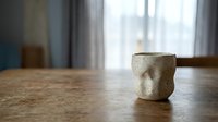 Handmade ceramic cup with rustic glaze sitting on wooden table near window with natural light, suitable for home decor and lifestyle content.